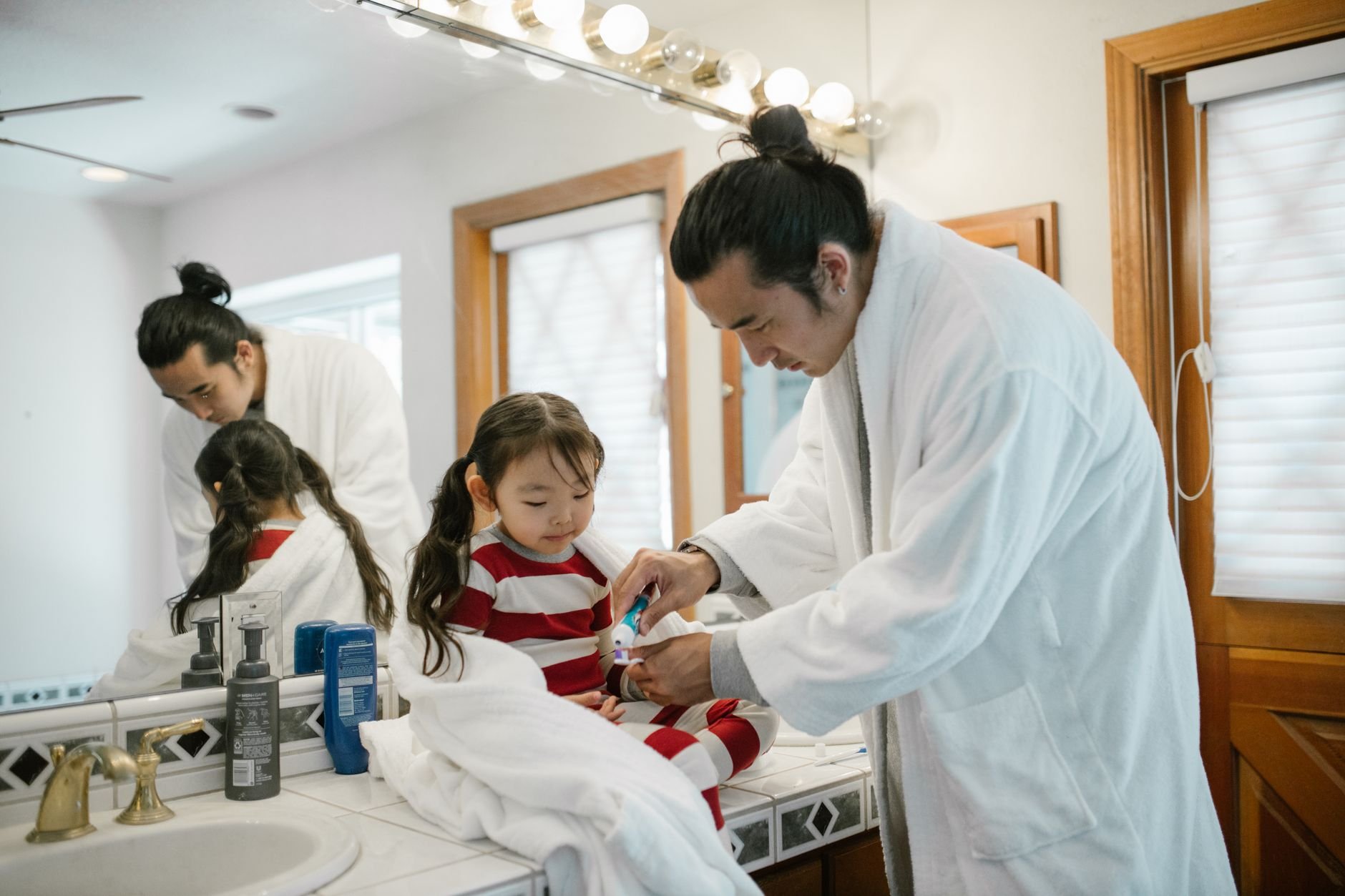 Father helping daughter brush teeth during a morning bathroom routine.